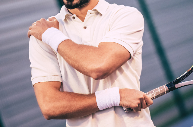 Un homme vêtu d'une chemise à col tient son épaule d'une main et une raquette de tennis de l'autre.