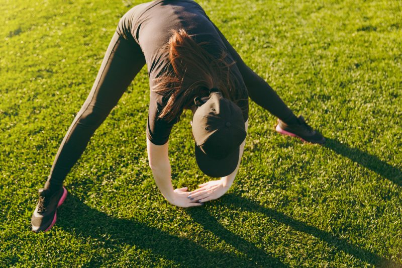 Jeune fille athlétique en uniforme noir, casquette faisant des exercices de sport, échauffement, étirement avant de courir sur la pelouse verte dans le parc du terrain de golf à l'extérieur le jour d'été ensoleillé.  Remise en forme, concept de mode de vie sain